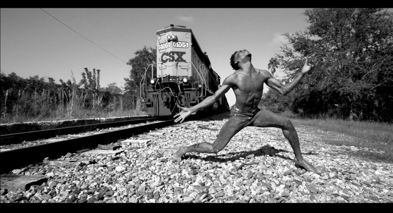 Beside a train track, Jared Brunson lowers into a kneel, his arms extended, his fingers splayed as a train approaches